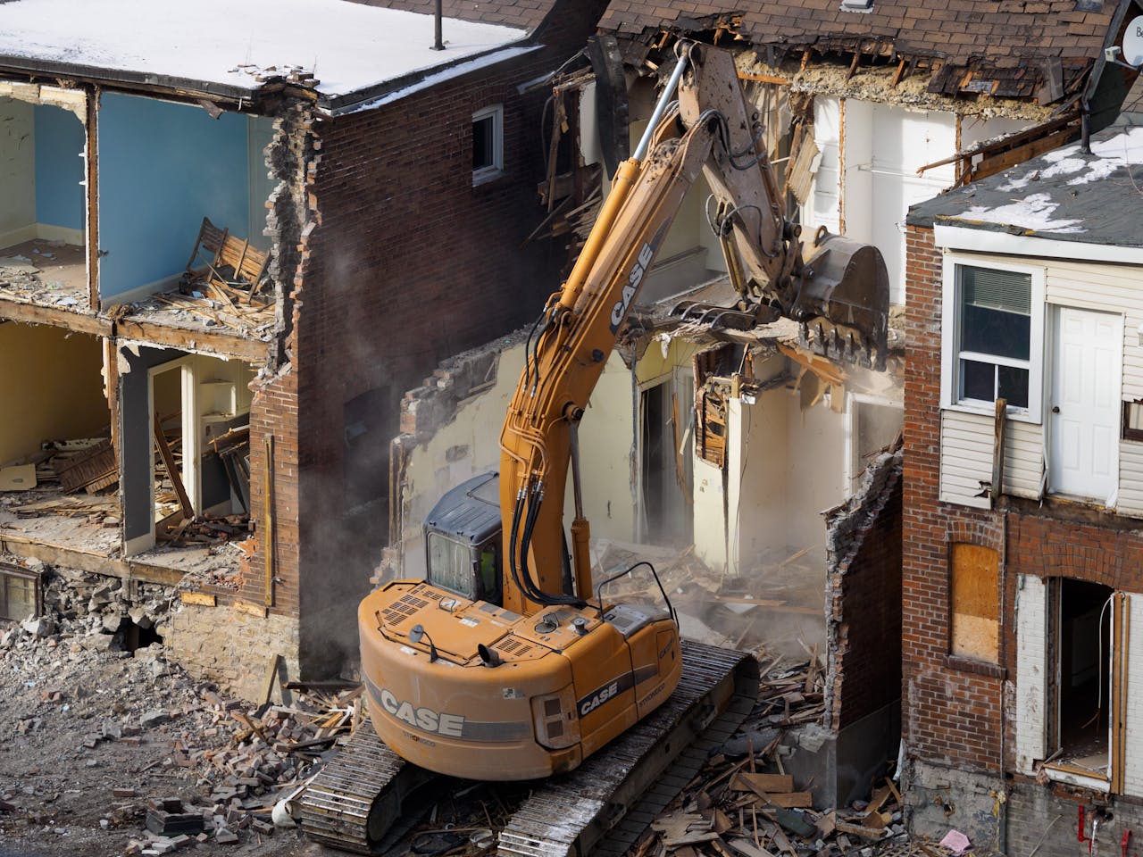 About An excavator demolishes an old brick building at a construction site, creating debris and dust.