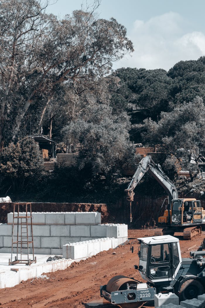 A busy construction site with heavy machinery, soil, and foliage around.